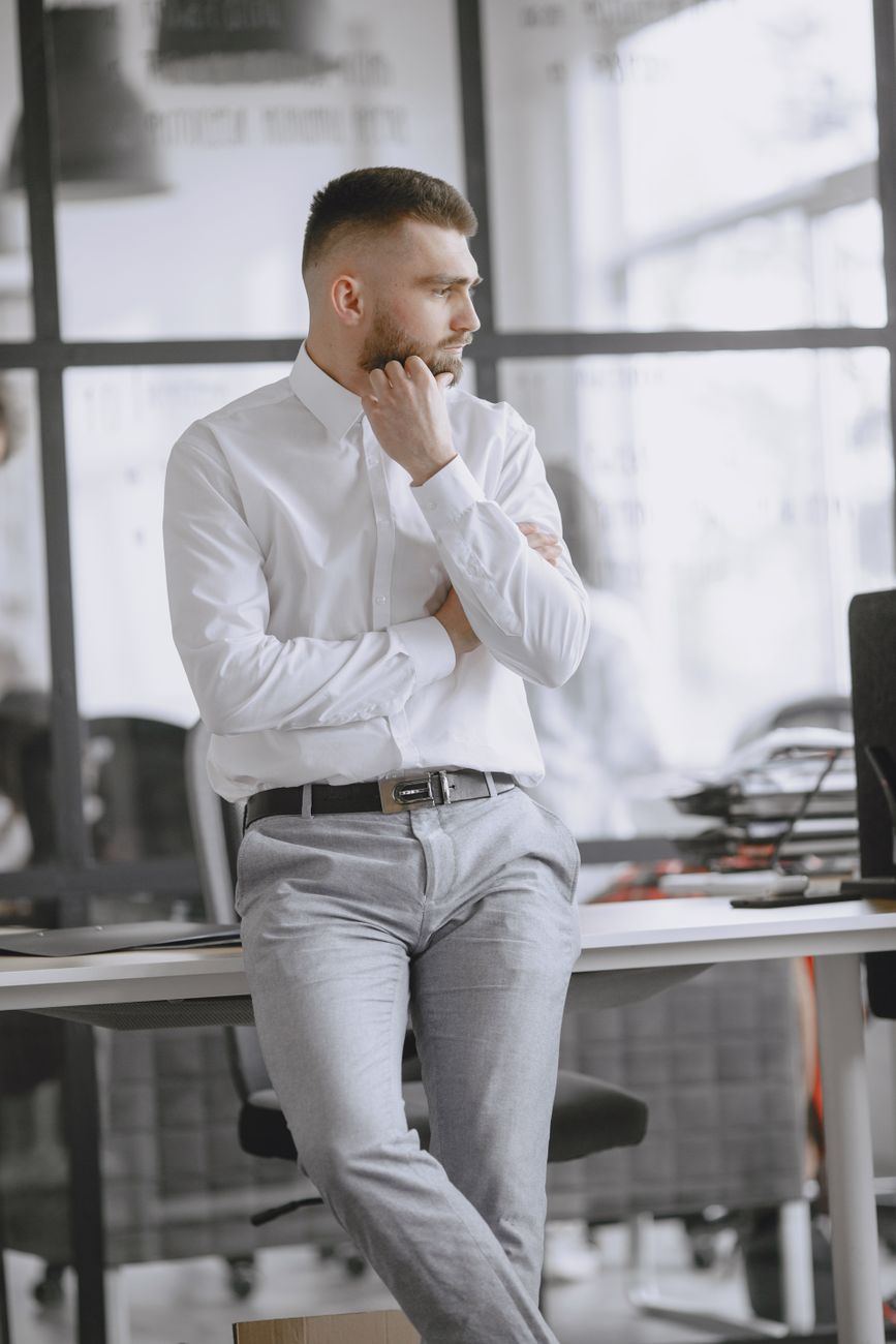 Homem de camisa apoiado numa mesa de escritório, olhando para o lado com a mão no queixo