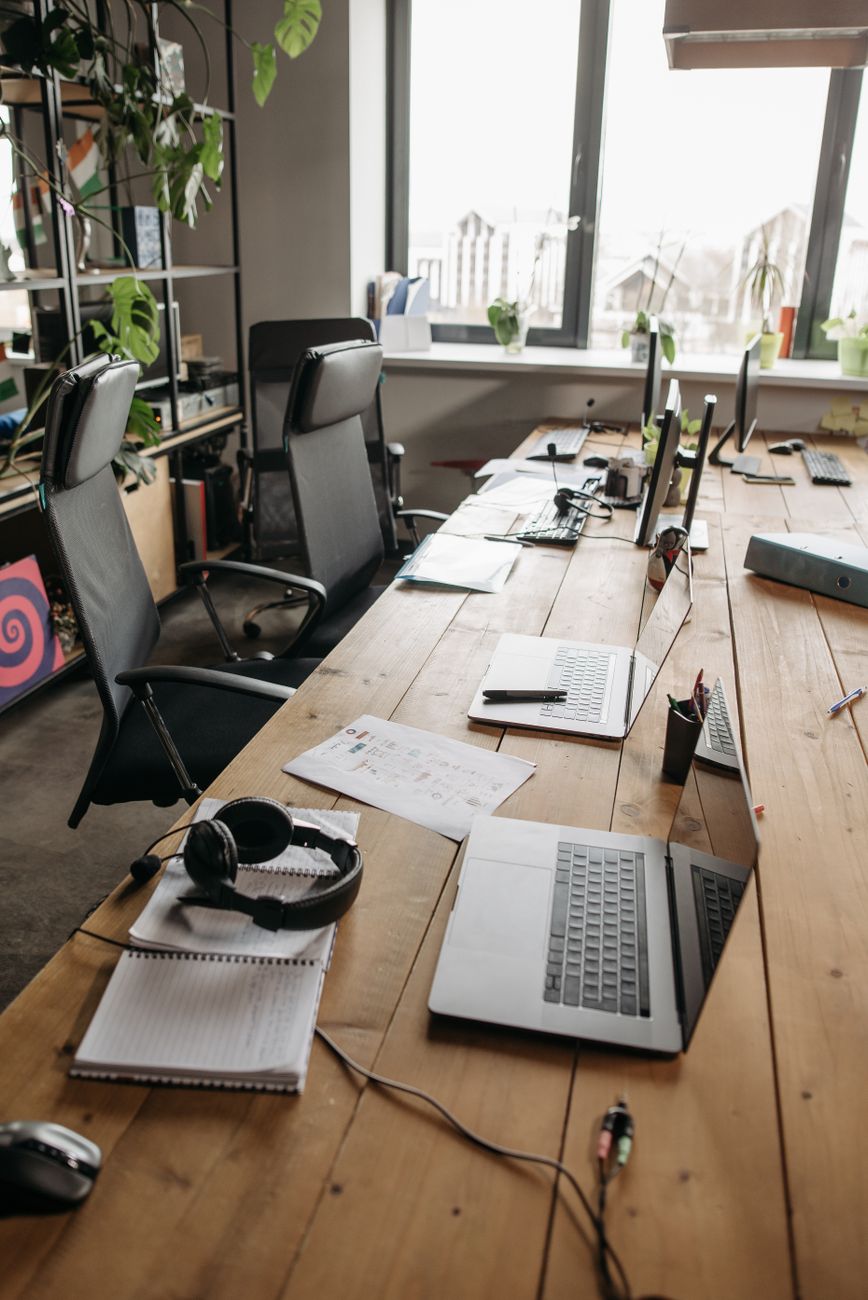 Mesa comprida de escritório compartilhada, com laptops, papéis, fones e cadeiras