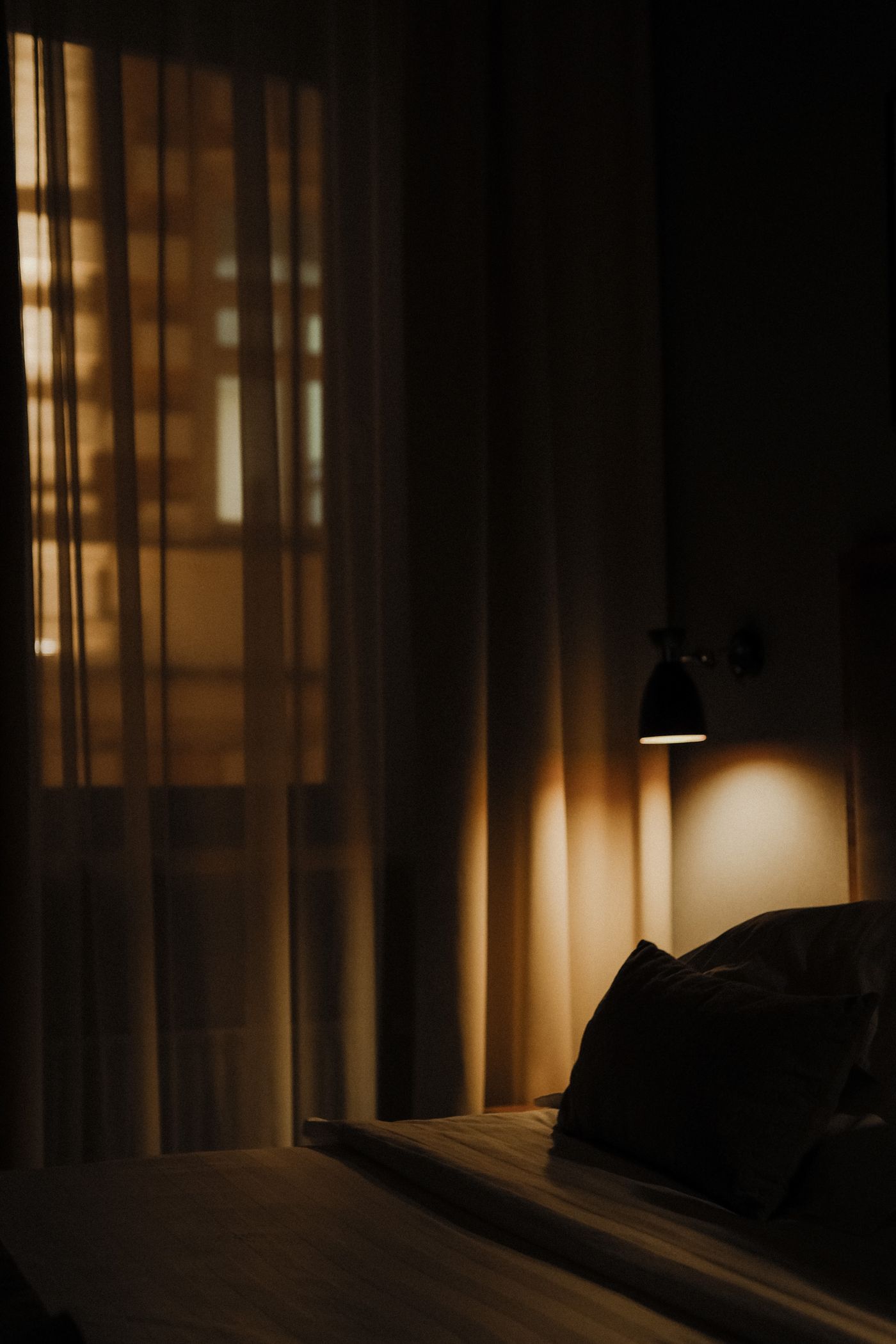 quarto à noite com luminária de leitura acesa junto à cabeceira e cortina translúcida