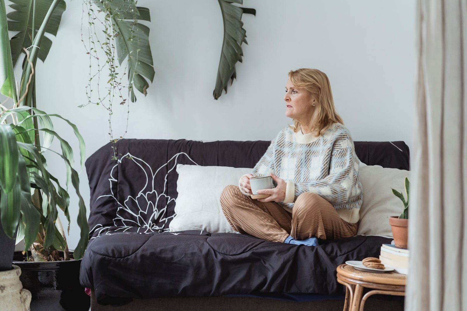 Mulher sentada no sofá da sala de casa, com uma xícara na mão, olhando o ambiente