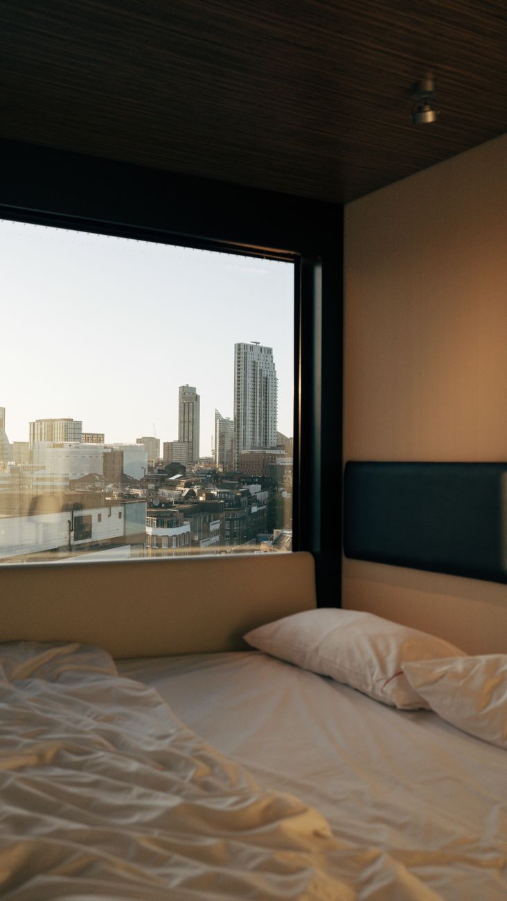 Cama desfeita junto de uma janela grande, com prédios da cidade do lado de fora