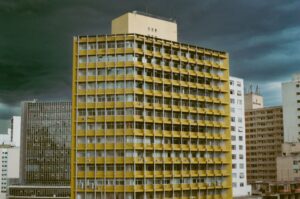 Prédio de apartamentos amarelo em São Paulo cercado por outros prédios, sob céu carregado