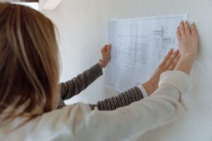 Mulher segurando uma planta baixa técnica aberta contra uma parede branca, vista por trás
